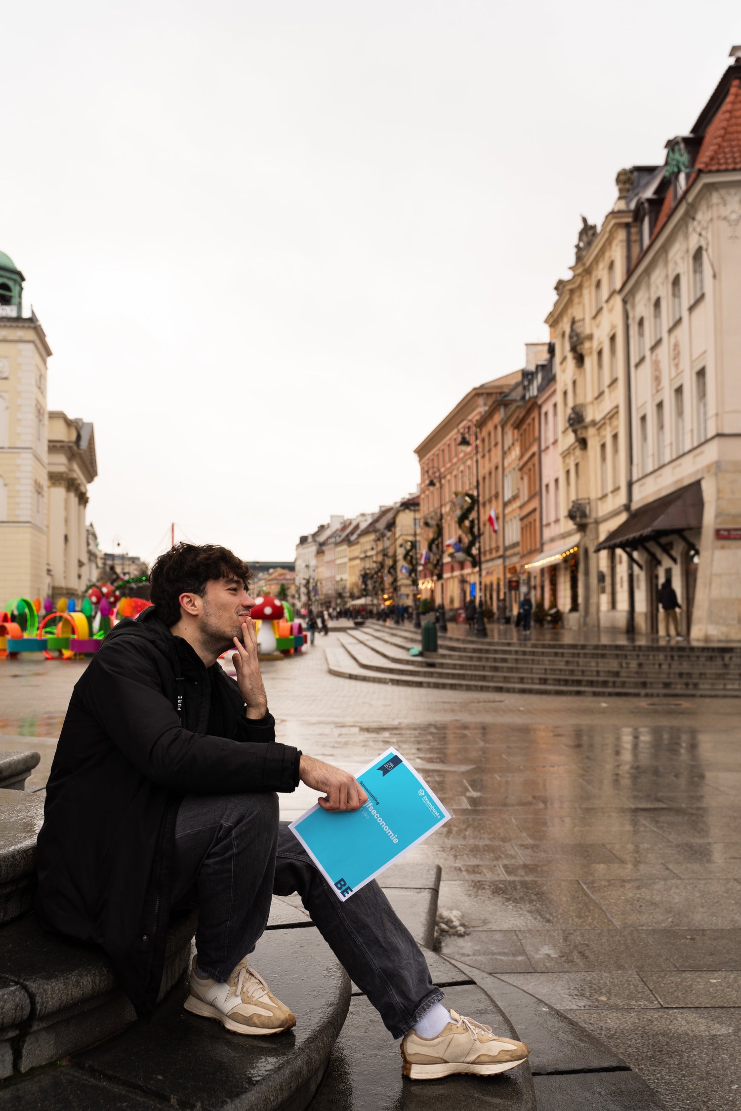 Student zit op een stenen trap in een Europese stad, verdiept in gedachten met een ExamBooks-studieboek in de hand, terwijl hij zich voorbereidt op zijn eindexamens.