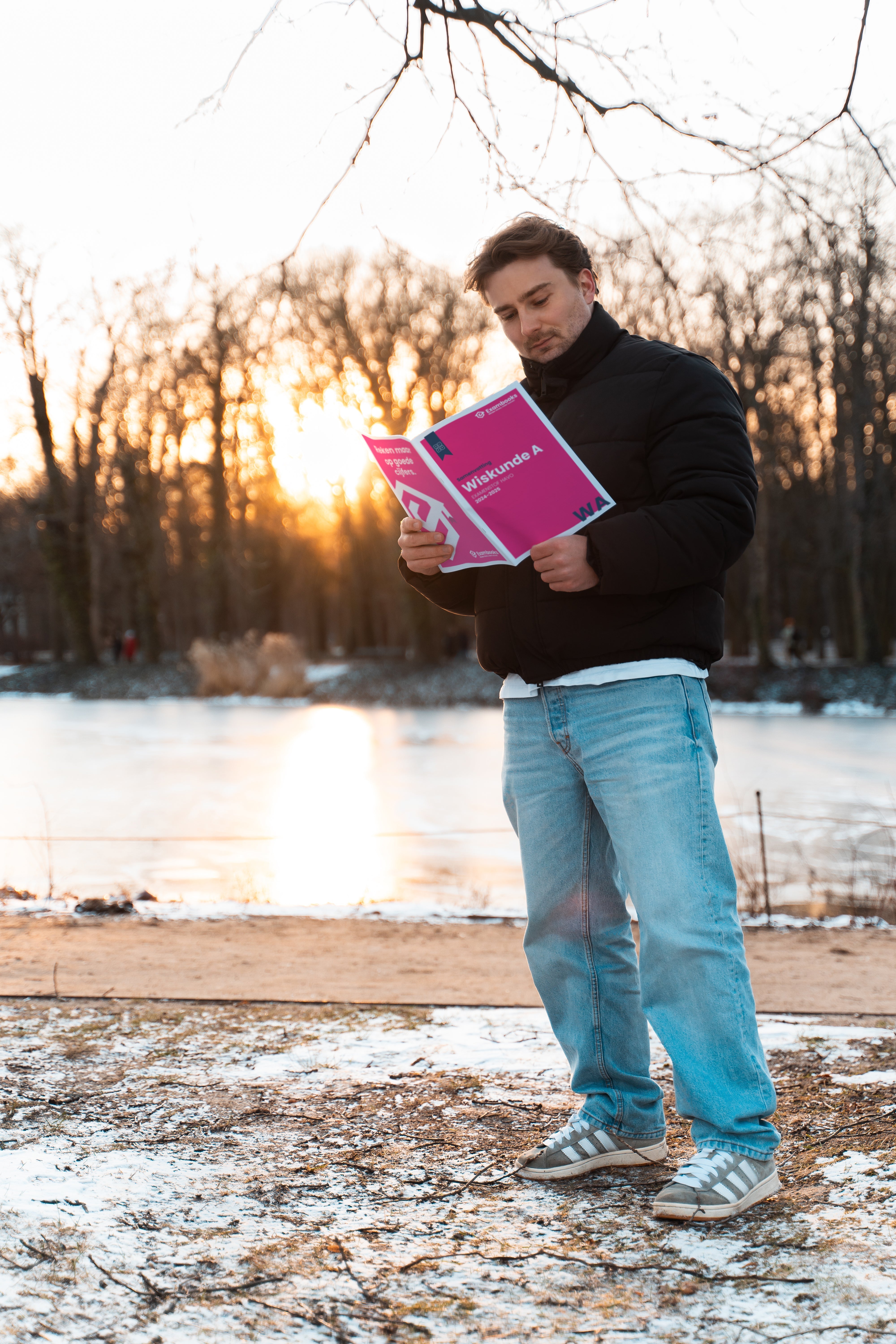 Student staat buiten in een winterse omgeving en leest geconcentreerd een ExamBooks-studieboek, terwijl hij zich voorbereidt op zijn eindexamens bij zonsondergang.