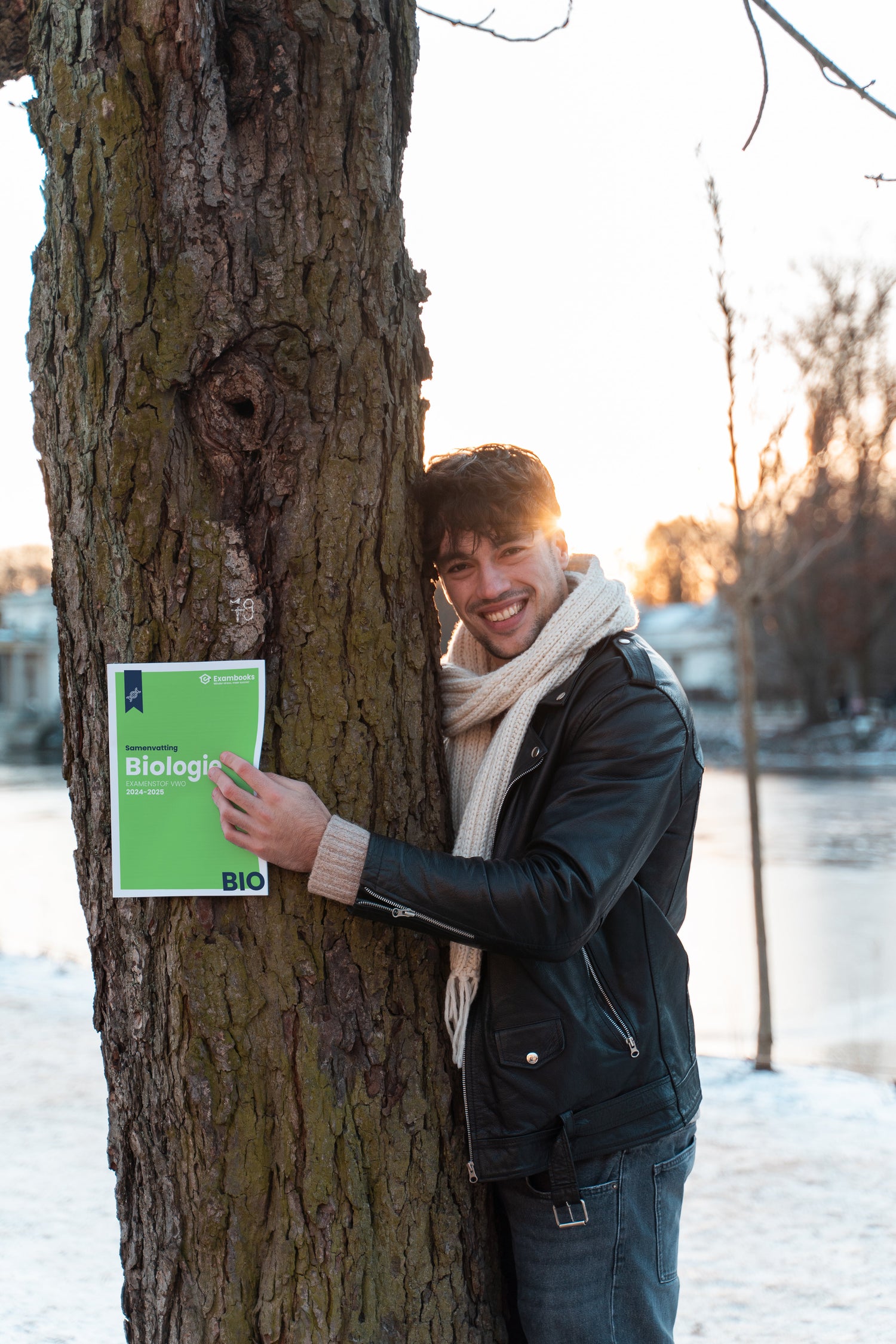 Blije student in winterkleding leunt tegen een boom terwijl hij een ExamBooks-biologieboek vasthoudt, enthousiast over zijn examenvoorbereiding in de buitenlucht.