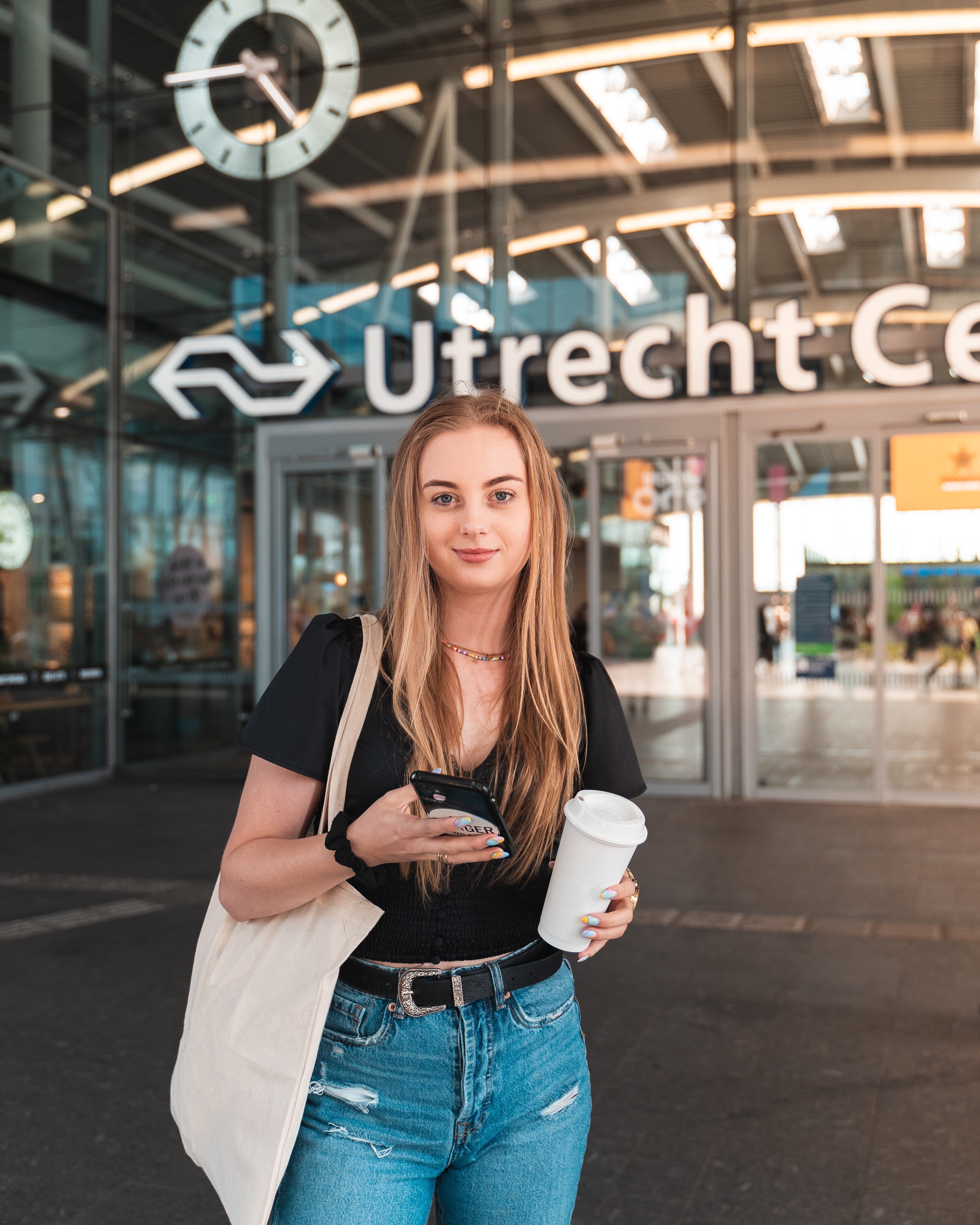 Jonge vrouw staat voor Utrecht Centraal met een koffiebeker en smartphone in de hand, klaar om te studeren met ExamBooks tijdens haar reis naar een rustige studieplek.